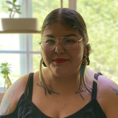 Image of Lupe Tejada Diaz wearing a black blouse and feather earrings. In the background is a window and plants